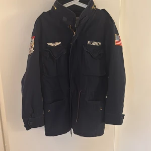Ralph Lauren Fieldjacka - Sällsynt mörkblå Field jacket från  Ralph Lauren med flera detaljer som broderade märken och amerikansk flagga på ärmen. Jackan har dragkedja och knappar framtill samt flera praktiska fickor. 10/10 skick. Strl: S/M Passar mig som är 182