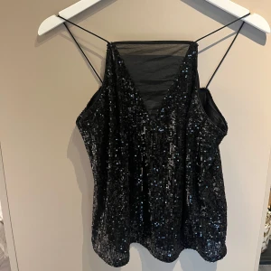 Svart paljettlinne - Snyggt svart linne med paljetter och tunna axelband. Perfekt för en kväll ute eller festliga tillfällen. Linnet har en v-ringad design och ger en glittrig look.