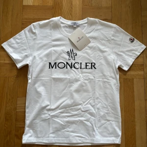 Vit t-shirt från Moncler - Säljer en stilren vit t-shirt från Moncler med deras ikoniska logga tryckt på bröstet och ett broderat emblem på ärmen. T-shirten har en klassisk passform och är tillverkad i mjuk bomull. Perfekt för en avslappnad och trendig look. Passar M/L