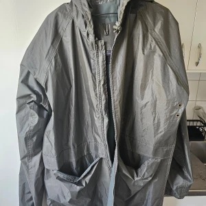 Grå regnjacka i nylon - Säljer en grå regnjacka i storlek XXL, tillverkad av 100% nylon. Jackan har en huva och dragkedja framtill, samt praktiska fickor. Perfekt för regniga dagar!