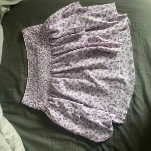 Lila blommig kjol - Charmig lila kjol med blommigt mönster. Kjolen har en volangdetalj och en elastisk midja för bekväm passform. Perfekt för en somrig look!