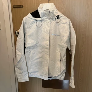 Vit vindjacka från Everest - Säljer en stilren vit vindjacka från Everest. Jackan har en praktisk huva och dragkedja framtill. Den är perfekt för blåsiga dagar och har en diskret logga på bröstet. Jackan är lätt och smidig att bära med sig.