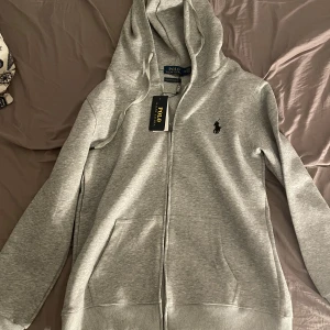 Grå hoodie från Ralph Lauren - Säljer en helt oanvänd stilren grå hoodie från Ralph Lauren med dragkedja och huva då den var för liten vid köp. Den har en broderad logga på bröstet och ribbade muddar vid ärmslut och nederkant. Perfekt för en avslappnad look.