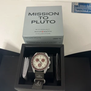Omega x Swatch Mission to Pluto  - Säljer en Omega x Swatch Mission to Pluto klocka från Bioceramic Moonswatch Collection. Klockan har en vit urtavla med röda detaljer och en grå textilrem. Den kommer i originalförpackning med ett extra gummi armband värt 800.