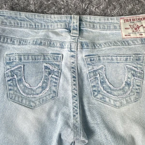 Ljusblå jeans från True Religion - Säljer ett par ljusblå jeans från True Religion med ikoniska bakfickor och broderad logga. De har en låg midja och en bootcut-stil, perfekt för en avslappnad look. Dragkedja och knapp framtill.