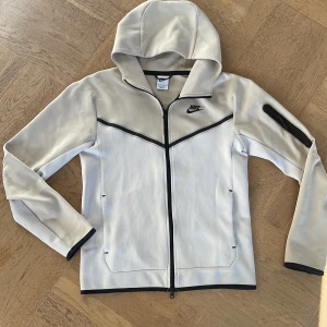 Beige nike tech zip up - Säljer en snygg beige hoodie från Nike med dragkedja och huva. Jackan har svarta detaljer och en ficka med dragkedja på ärmen. Perfekt för en sportig look eller en avslappnad dag. 🖤