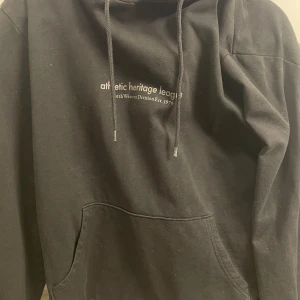Svart hoodie med tryck - Säljer en svart hoodie med texten 'athletic heritage league' på bröstet. Den har en klassisk design med dragsko i huvan och en stor magficka. Perfekt för en avslappnad stil.