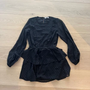 Svart klänning med volangdetaljer - Snygg svart klänning med volangdetaljer och diskret prickigt mönster. Klänningen har långa ärmar och en peplumliknande design i midjan. Perfekt för en stilren look.