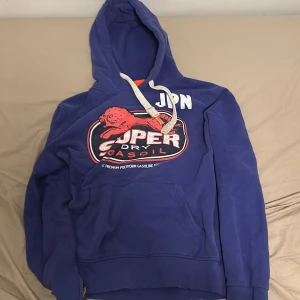 Blå hoodie från Superdry - Säljer en blå hoodie från Superdry med tryck på framsidan. Den har en justerbar huva med vita snören och en stor ficka framtill. Perfekt för en avslappnad stil.