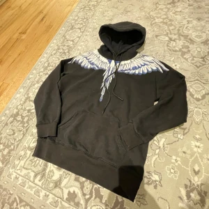 Marcelo Burlon Hoodie - Säljer en svart hoodie från Marcelo Burlon med ett iögonfallande vitt och blått vingmönster över axlarna. Tröjan har en klassisk känguruficka och justerbar huva med snören. Perfekt för en stilren och unik look.