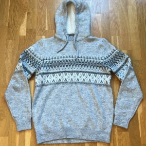 Grå stickad hoodie med mönster - Säljer en mysig grå stickad hoodie med ett snyggt mönster i svart och vitt över bröstet. Tröjan har långa ärmar och en praktisk huva med dragsko. Perfekt för kyliga dagar!