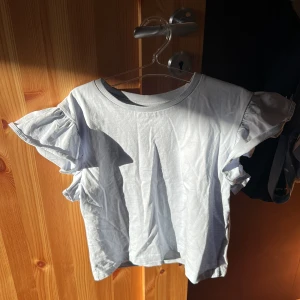 Ljusblå topp med volangärmar från Kappahl - Säljer en söt vit topp från Kappahl med volangdetaljer på ärmarna 💕. Perfekt för en somrig look. Toppen har en rund halsringning och är tillverkad i ett mjukt material. Har bara använt ett fåtal gånger. Säljer på grund av att den är för liten!💗