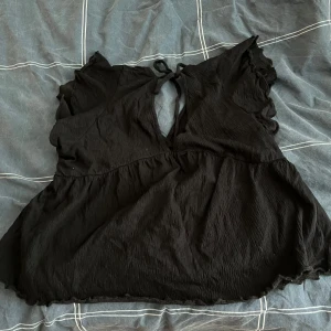 Svart blus med knytband - Säljer en svart blus med v-ringning och knytband i nacken. Blusen har volangdetaljer vid ärmslut och nederkant, vilket ger en romantisk touch. Perfekt för en stilren look.