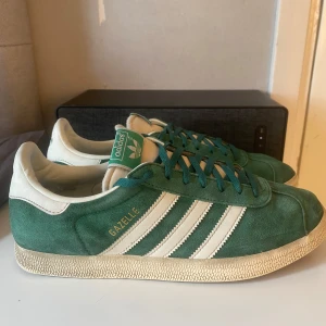 Gröna Adidas Gazelle sneakers - Snygga gröna Adidas Gazelle sneakers med vita detaljer och klassiska tre ränder på sidan. Skorna har en ovandel i mocka och snörning framtill. Perfekta för en stilren och sportig look.                                  OBS sko box finns inte!