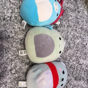 Squishmallows  - Tre söta Squishmallows mjukisdjur i olika färger och former. En ljusblå med röd rosett, en ljusgrön sköldpadda och en ljusgrå med röd halsduk. Perfekta för kramar och mysiga stunder!