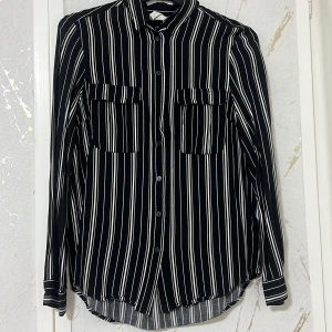 Svartvit randig skjorta - Snygg svartvit randig skjorta med långa ärmar och knappar framtill. Skjortan har två bröstfickor och en klassisk krage. Perfekt för en stilren look.