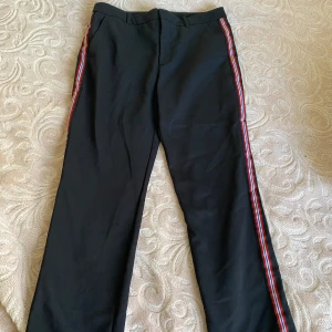 Svarta byxor med ränder från Zara Basic - Snygga svarta byxor från Zara Basic med färgglada ränder längs sidorna. Byxorna har en dragkedja och knapp framtill för enkel stängning. Perfekta för en stilren look med en twist. Priset går alltid att diskutera💞