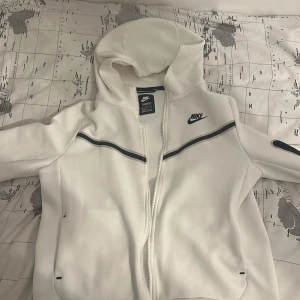 Vit huvtröja från Nike - Säljer en stilren vit huvtröja från Nike med dragkedja och svarta detaljer. Tröjan har en justerbar huva och långa ärmar med en praktisk ficka på ärmen. Perfekt för en sportig look eller avslappnade dagar.