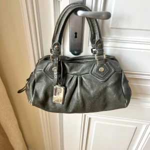 Marc by Marc Jacobs väska - Marc Jacobs väska i modellen Classic Q! Grå aktig, med coola vintage detaljer! Väskan är i bra skick, finns täcken på användning som syns i bilderna! Fråga gärna om ni undrar något! Kan mötas upp i Stockholm eller frakta, tryck gärna köp direkt!