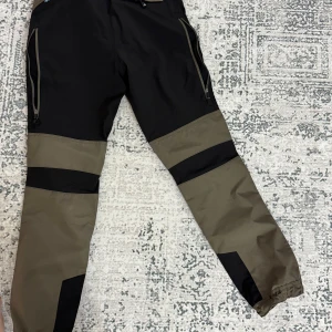 Grön och svart byxa från Arrak Outdoor - Säljer en grön och svart byxa från Arrak Outdoor. Byxan har flera praktiska fickor med dragkedja och förstärkta knäpartier. Perfekt för utomhusaktiviteter med justerbara benslut för optimal passform.