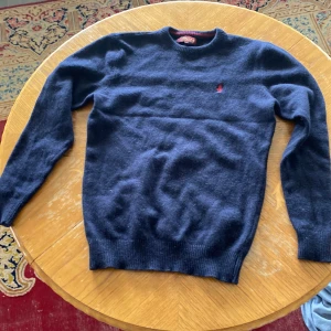 Mörkblå ulltröja från MCS - Säljer en snygg mörkblå ulltröja från MCS med ett rött broderat emblem på bröstet. Tröjan har långa ärmar och ribbade muddar vid ärmslut och nederkant. Perfekt för kyliga dagar!