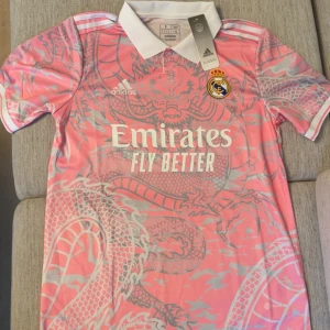 Real Madrid rosa fotbollströja Adidas - Snygg rosa Real Madrid fotbollströja från Adidas med vit krage och korta ärmar. Tröjan har ett stort drakmönster i silvergrått över hela framsidan och baksidan, samt klubbmärke och Adidas-logga på bröstet. Materialet är lätt och andas, perfekt för träning eller match.