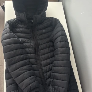 Svart pufferjacka från 156FUNK - Svart pufferjacka från 156FUNK med huva och horisontella sömmar. Jackan har dragkedja framtill och är tillverkad i syntetmaterial. Perfekt för kalla dagar när du vill hålla värmen och ändå se cool ut.