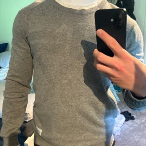 Grå stickad tröja Armani Exchange - Snygg grå stickad tröja från Armani Exchange med diskret logga på bröstet och patch på nederkanten. Rund halsringning med vit detalj och långa ärmar. Perfekt för dig som gillar stilren och enkel design.