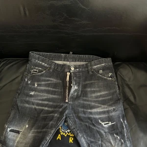 Svarta Dsquared2 jeansshorts med slitningar - Snygga svarta jeansshorts från Dsquared2 med slitna detaljer och en stor vit patch med text bak på linningen. Jeansen har dragkedja framtill, klassisk femficksdesign och en cool, tvättad look. Perfekta för dig som gillar streetstyle och statement-plagg.