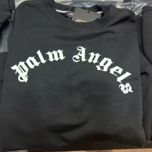 Svart Palm Angels sweatshirt - Svart sweatshirt från Palm Angels med klassisk vit logotyp i gotisk stil över bröstet. Tröjan har rund halsringning och långa ärmar. Tillverkad i mjuk bomull som känns skön mot huden. Perfekt för dig som gillar streetwear och vill sticka ut. Finns i alla storlekar 