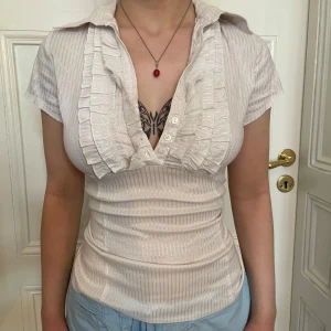 Vit v-ringad skjorta med volang - Snygg vit skjorta med v-ringning och volangdetaljer framtill. Skjortan har korta ärmar, knappar och ett diskret randigt mönster i tyget. Figurnära passform som framhäver siluetten och ger en fräsch look.