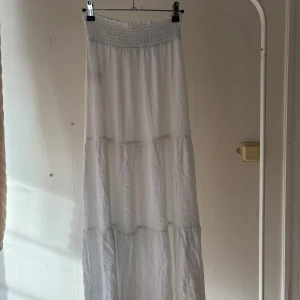 Vit långkjol med smock i midjan - Säljer en flowy vit långkjol med bred smock i midjan. Kjolen har flera volanger och är gjord i ett tunt, lätt tyg som ger en luftig känsla. Perfekt för varma dagar och enkel att styla med crop tops eller linnen.