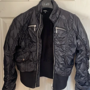 Svart bomberjacka från DKNY - Säljer en svart croppad bomberjacka från DKNY med glansig yta, ribbade muddar och dragkedjor både framtill och på bröstfickorna. Jackan har coola rynkade detaljer och mjukt foder på insidan. Perfekt för dig som vill ha en streetig och snygg look.