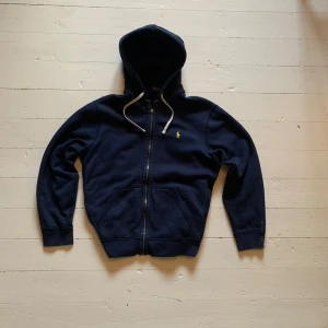 Hoodie Polo Ralph Lauren marinblå - Snyyygg hoodie från Ralph Lauren, den är marinblå med gul broderad logga och vita snören. Förutom att själva grejen man håller i på dragkedjan har gått av (se bild 5) så är den i mycket bra skick, skulle säga att den sitter som en storlek M/L!