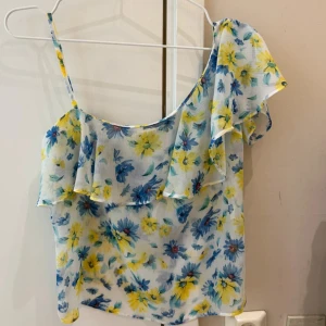 Blommig volangtop med smala axelband - Superfin topp med blommigt mönster i blått och gult på vit botten. Tunt, luftigt material och volangdetalj framtill samt över ena axeln. Ena axelbandet är smalt och den andra sidan har en bred volangärm för asymmetrisk look. Perfekt för varma dagar.