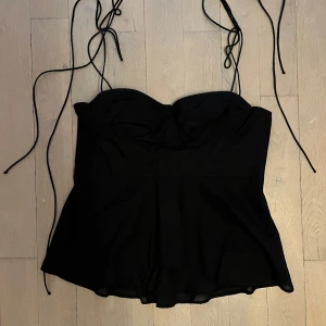 Svart korsettopp från Zara - Supersnygg svart korsettopp från Zara med tunna knytband och lätt utsvängd nederdel. Toppen har vadderade kupor och smock i ryggen för extra komfort. Perfekt att styla med jeans eller kjol för en trendig look.