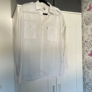 Vit skjorta från Bogart i bomull - Klassisk vit skjorta från Bogart i storlek M. Skjortan har två bröstfickor med knappar, axelklaffar och långa ärmar. Tillverkad i bomull för en skön och luftig känsla. Perfekt för dig som gillar en clean och stilren look.
