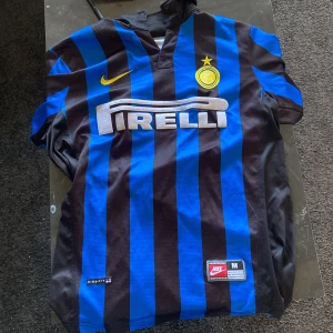 Inter Ronaldo 7 Nike fotbollströja M - Klassisk Inter Milan fotbollströja med blå och svarta ränder, Pirelli-tryck på bröstet och Ronaldo 7 på ryggen. Tröjan har korta ärmar, gul Nike-logga och Inter-emblem. Tillverkad i lätt och ventilerande polyester, perfekt för fotbollsälskare.