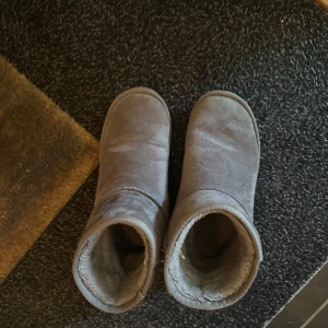 Grå mockaboots med foder - Mysiga grå boots i mocka med mjukt foder på insidan. Skorna har rund tå och platt sula, perfekta för kalla dagar. Klassisk enkel design som passar till det mesta och håller fötterna varma hela vintern.