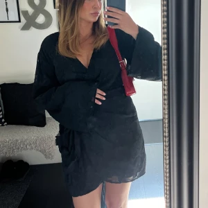 Omlottklänning  - Snygg svart kort omlottklänning med knyt i midjan och vida långa ärmar. Fina detaljer med blommor 🖤klänningen är endast använd en gång! 