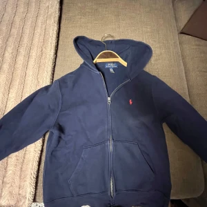 Mörkblå hoodie från Polo Ralph Lauren - Snygg mörkblå hoodie från Polo Ralph Lauren med dragkedja och huva. Klassisk röd broderad logga på bröstet och stora fickor framtill. Tillverkad i mjuk bomull, perfekt för chill dagar. Passar dig som gillar stilren och enkel streetwear.