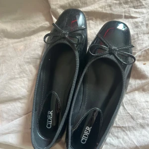 Svarta ballerinaskor från Cider - helt nya: Glansiga svarta ballerinaskor från Cider med rund tå och söt rosettdetalj framtill. Skorna har en klassisk och stilren look som passar perfekt till kjol eller klänning. Platt sula och mjukt material för bekväm känsla. 