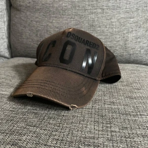 Brun Dsquared2 ICON keps - Snygg brun keps från Dsquared2 med stor svart 'ICON'-text framtill och justerbart spänne bak. Klassisk böjd skärm och broderad text 'DEAN & DAN CATEN' baktill. Tillverkad i bomull, perfekt för en cool streetstyle-look.
