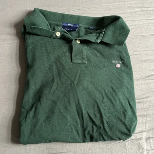 Grön pikétröja från GANT - Klassisk grön pikétröja från GANT med broderad logga och sköld på bröstet. Tröjan har korta ärmar, krage och två knappar framtill. Tillverkad i mjuk bomull som känns skön mot huden. Perfekt för en avslappnad och stilren look. Hör av er vid minsta lilla fråga 