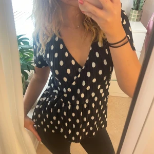 Svart prickig omlottblus/topp - Supersöt svart omlottblus/topp med vita prickar och korta ärmar. Blusen har v-ringning, plisserat tyg och markerad midja som ger en snygg siluett. Perfekt för dig som gillar lekfulla mönster och trendiga detaljer. 