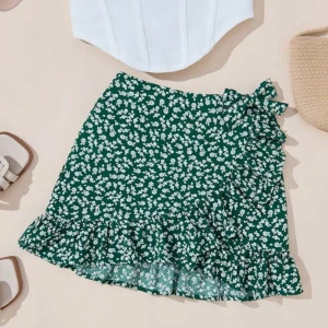 Grön blommig volangkjol med knyt - Supersöt grön kortkjol med vitt blommönster, volangkant och knyt i sidan. Perfekt flowy känsla och lätt material som passar till crop tops eller linnen. Kjolen har omlott-look och ger en fräsch vibe till din outfit.