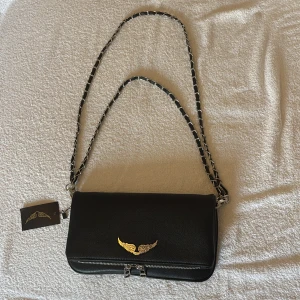 Svart axelväska Zadig & Voltaire skinn - Snygg svart axelväska i skinn från Zadig & Voltaire med kedjerem och läderdetaljer. Väskan har ett silverfärgat vingemblem framtill och dubbla dragkedjor nertill. Köpt för 3600kr men inte den modellen jag ville ha då jag fick den som present💞