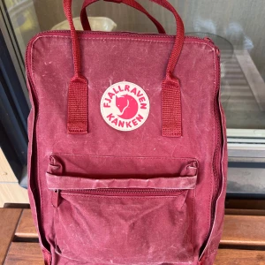 Röd Fjällräven Kånken Laptop 15” ryggsäck - Klassisk röd Fjällräven Kånken ryggsäck med ikonisk rund logotyp framtill. Ryggsäcken har ett stort huvudfack, framficka med dragkedja och justerbara axelremmar. Tillverkad i slitstark polyester och har praktiska handtag upptill. Väskan är hel. Sittdyna medföljer ej. Nypris 1699:-
