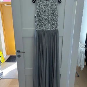 Lång grå klänning med paljetter - Supersnygg långklänning i grått med glittrig paljettöverdel och plisserad kjol i tunt tyg. Ärmlös modell med rund halsringning och dragkedja bak. Perfekt för bal eller fest.