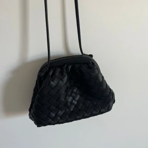 Svart flätad clutchbag  - Snygg svart axelväska från BOW19 med flätad design i skinnimitation. Väskan har en mjuk, rundad form och stängs med metallspänne. Invändigt finns ett fack med dragkedja för småsaker. Den är i helt nyskick endast använd fåtal gånger, väldigt fin❤️Snörena på väskan är justerbara så man kan göra de så långa/korta som man vill!